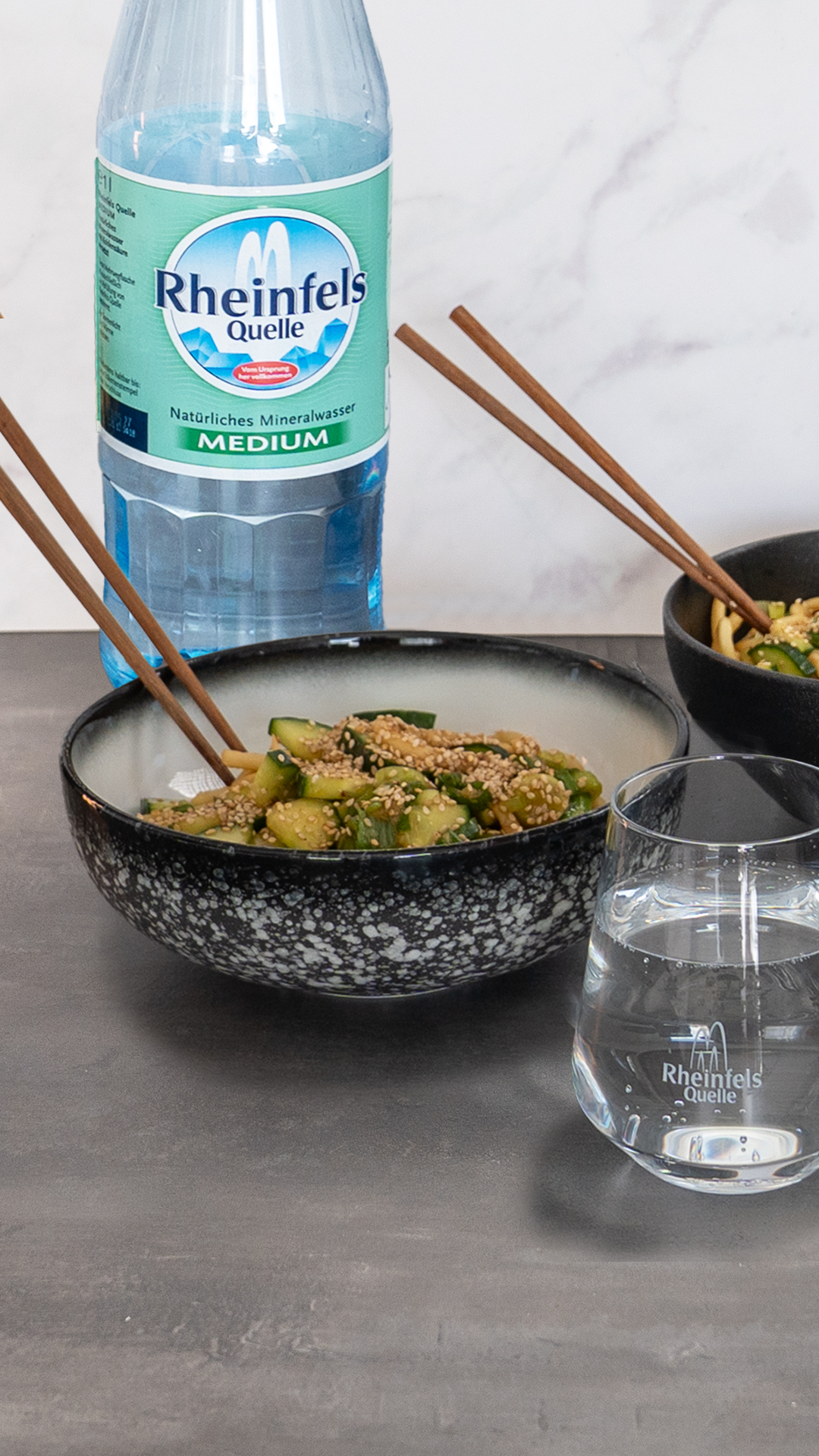 Spicy Udon mit Gurke und Sesam mit einer Flasche Rheinfels Quelle Medium auf einem Holzbrett in Schüssel serviert