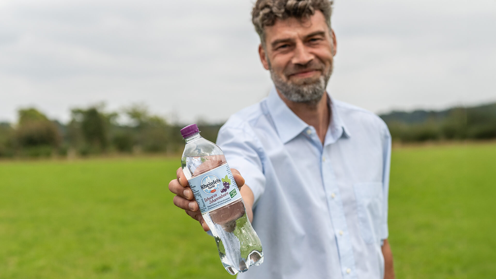 Mann hält lächelnd eine Flasche Schwarze Johannisbeere in die Kamera.