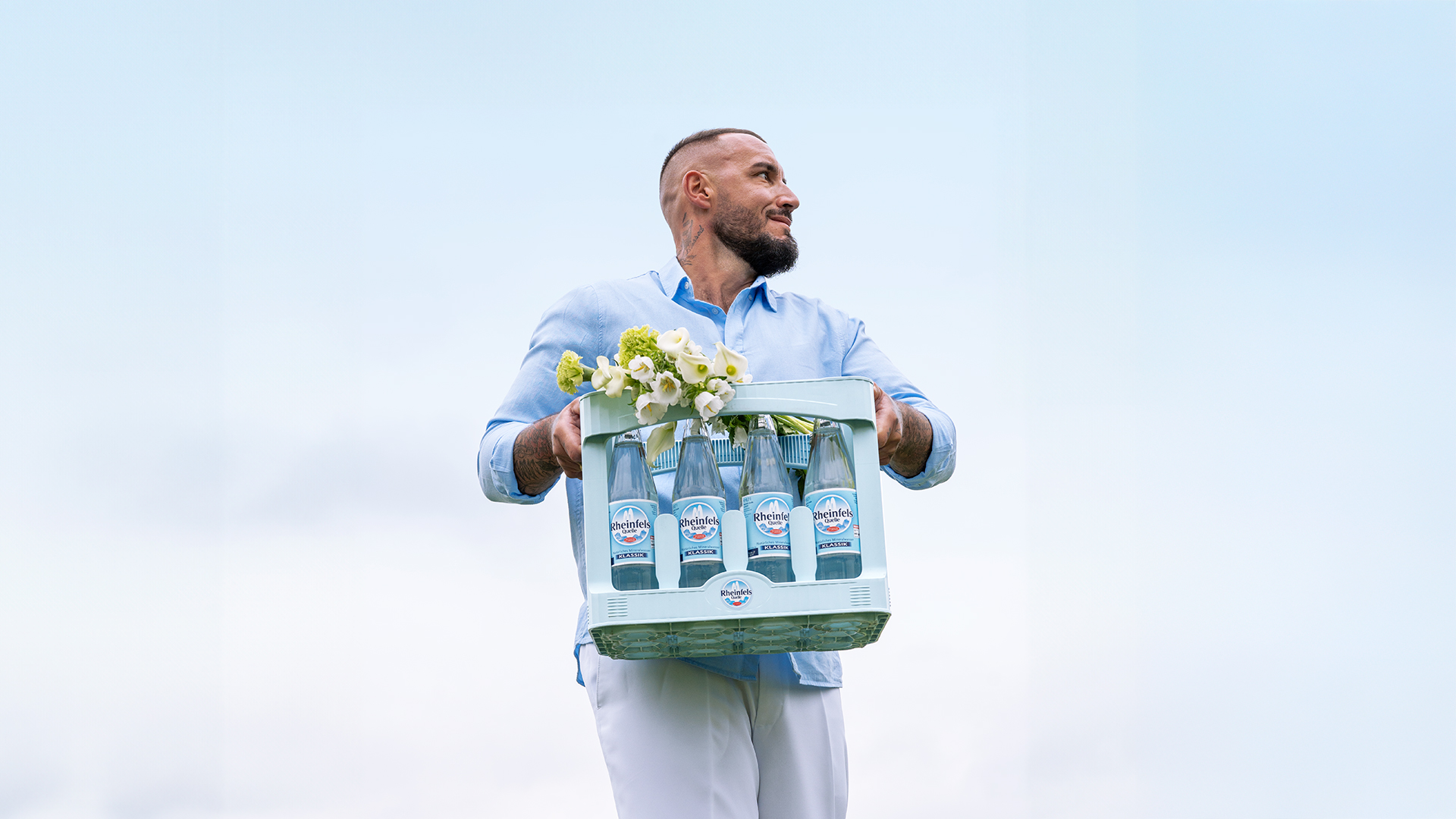 Mann läuft über eine Wiese auf dem Weg zum Dinner. Er trägt einen Kasten Rheinfels Quelle Klassik.